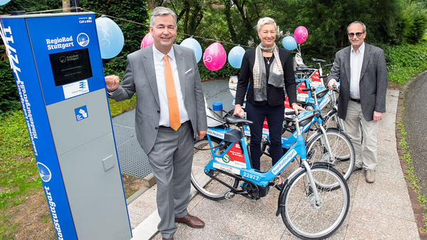 Trois personnes se tiennent près de vélos en libre-service. Un homme en costume et une femme debout sur un vélo sourient, tandis qu'un autre homme en lunettes est à côté. En arrière-plan, des ballons colorés ajoutent une ambiance festive, près d'une station de location de vélos.