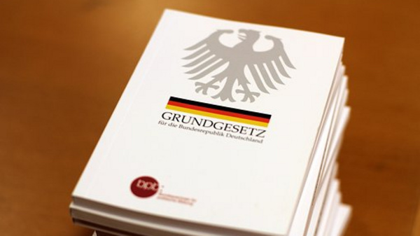A stack of white-covered books featuring a double-headed eagle emblem. The top book displays the title "GRUNDGESETZ für die Bundesrepublik Deutschland" in bold letters, along with a horizontal black, red, and yellow stripe beneath the title, representing the German flag.