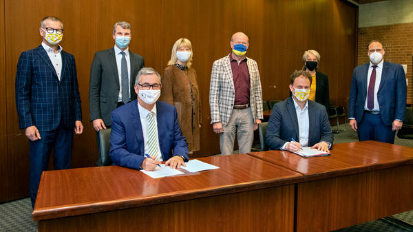 Eight individuals are gathered in a formal setting, wearing masks. Two men at a table in the foreground are signing documents, while others stand behind them, observing the event. The scene depicts a moment of collaboration or agreement among the group.