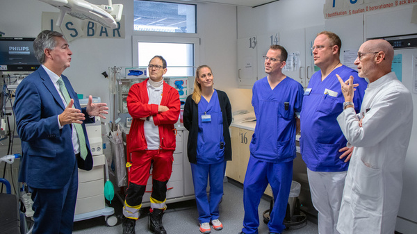 Ein Mann im Anzug spricht zu einer Gruppe von sechs Personen in medizinischer Kleidung, darunter ein Sanitäter und mehrere Ärzte, in einem Krankenhausraum. Die Gruppe hört aufmerksam zu, während sie in einem Gespräch vertieft ist.
