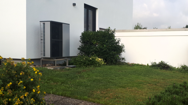 Eine moderne Wärmepumpe steht neben einem Haus auf einer Rasenfläche. Im Vordergrund sind gelbe Blumen und Sträucher zu sehen. Im Hintergrund erkannte man einen niedrigen Blick auf eine weiße Wand, die den Garten umschließt. Der Himmel ist leicht bewölkt.