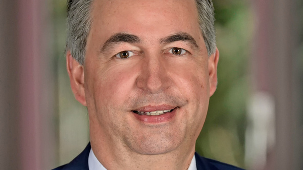 A man with short, light gray hair smiles warmly. He is dressed in a dark suit and a white shirt with a pink striped tie. The background features soft, blurred greenery, suggesting an outdoor setting.