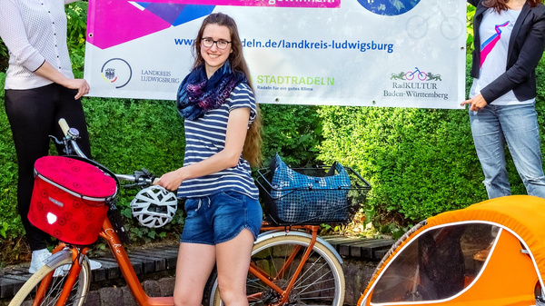 Drei Frauen stehen vor einem Banner, das zur Teilnahme an einer Radveranstaltung namens "STADTRADELN" einlädt. Eine Frau posiert mit einem Fahrrad und einem Anhänger, während die anderen beiden das Banner halten, das Informationen und die Aufforderung zur Anmeldung enthält.