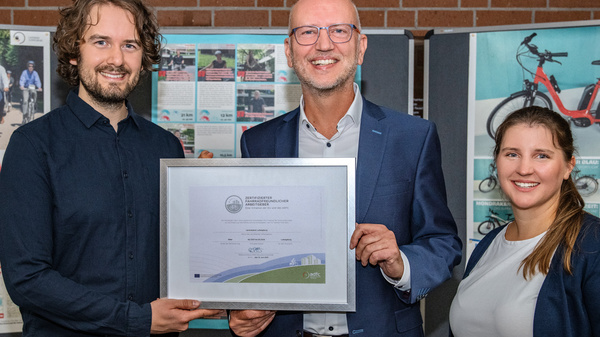 Drei Personen stehen zusammen und halten eine Urkunde in der Mitte. Im Hintergrund befinden sich Plakate mit Informationen zum Radfahren zur Arbeit. Die Person links hat lockiges Haar, die mittlere Person trägt eine Brille, und die rechts hat lange Haare.