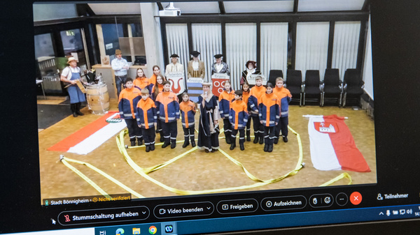 Auf einem Laptop-Bildschirm sind Kinder in orangefarbenen Uniformen zu sehen, die in einer Gruppenaufstellung sitzen. Im Hintergrund sind Fahnen und eine Videokonferenz mit weiteren Teilnehmern sichtbar. Die Szene vermittelt ein Gefühl der Gemeinschaft und des Engagements.