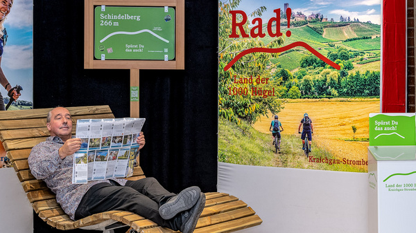 Eine Ausstellung mit dem Thema Outdoor-Aktivitäten zeigt zwei großflächige Plakate: eines mit der Aufschrift „Zu Fuß!“ und das andere „Per Rad!“. In der Mitte sitzt ein Mann auf einer Liege und betrachtet eine Reisebroschüre. Im Hintergrund sind Angebote für Wander- und Radfahrtouren dargestellt.
