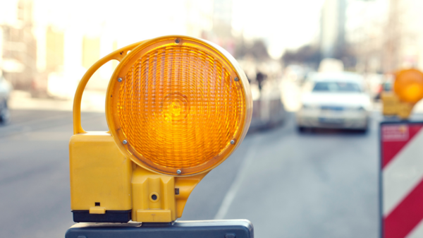Ein gelbes Warnlicht ist auf einem Verkehrszeichen montiert, das durch diagonale rote und weiße Streifen gekennzeichnet ist. Im Hintergrund sind unscharfe Autos auf einer Straße sichtbar, was darauf hinweist, dass sich die Szene in einem städtischen Umfeld befindet.