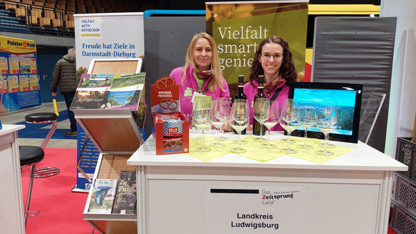 Deux femmes se tiennent derrière un stand d'exposition avec des verres à vin au premier plan. Le stand représente le Landkreis Ludwigsburg, et des brochures sont visibles sur la table, affichant des informations sur la région. Une ambiance accueillante et informative se dégage de l'espace d'exposition.