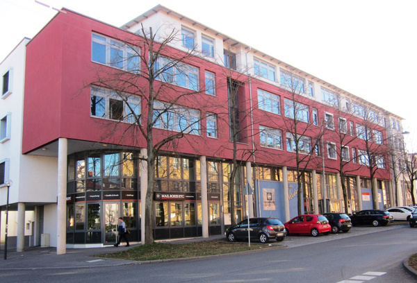 Ein modernes, rotes Gebäude mit großen Fenstern und einer Glasfassade im Erdgeschoss. Vor dem Gebäude sind parkende Autos und einige Bäume zu sehen. Der Gehweg führt entlang des Gebäudes, auf dem ein Passant geht.
