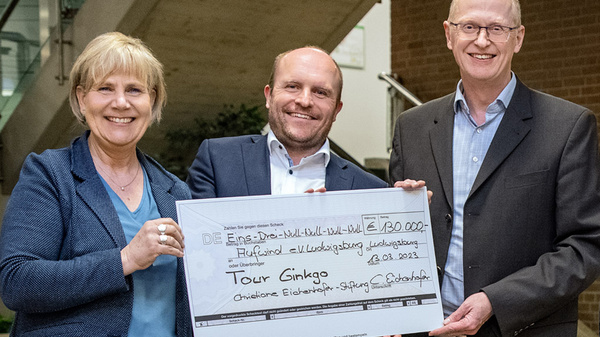 Drei Personen stehen zusammen und halten einen großen Scheck über 130.000 Euro für das Projekt "Tour Ginkgo". Die Gesichter zeigen Freude und Stolz. Die Veranstaltung findet am 23.03.2023 statt.