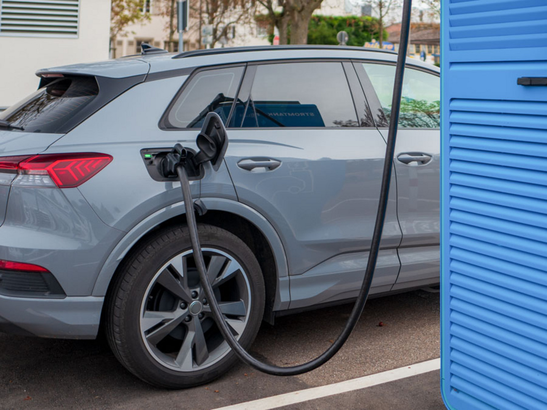 Ein graues Elektroauto steht an einer Ladestation. Ein Ladekabel ist in die Fahrzeuganschlussdose eingesteckt. Die Umgebung zeigt eine moderne Stadtlandschaft mit mehreren Gebäuden im Hintergrund. Die Szene vermittelt den Akt des Ladens eines Elektrofahrzeugs.