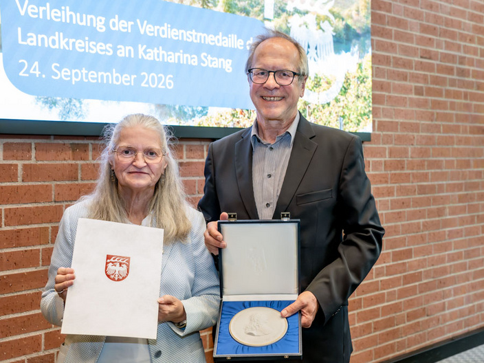 Eine Frau hält ein Zertifikat in der Hand, während ein Mann eine Auszeichnung in einer Box präsentiert. Beide lächeln vor einer von Bäumen umgebenen Wand mit einer Tafel, die den Anlass der Verleihung anzeigt. (Text KI generiert)