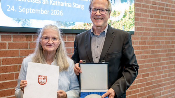 Eine Frau hält ein Zertifikat in der Hand, während ein Mann eine Auszeichnung in einer Box präsentiert. Beide lächeln vor einer von Bäumen umgebenen Wand mit einer Tafel, die den Anlass der Verleihung anzeigt. (Text KI generiert)