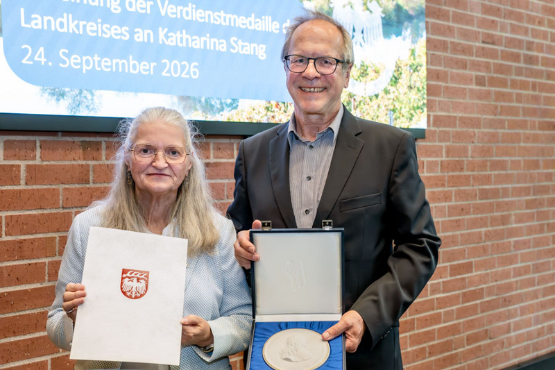 Eine Frau hält ein Zertifikat in der Hand, während ein Mann eine Auszeichnung in einer Box präsentiert. Beide lächeln vor einer von Bäumen umgebenen Wand mit einer Tafel, die den Anlass der Verleihung anzeigt. (Text KI generiert)