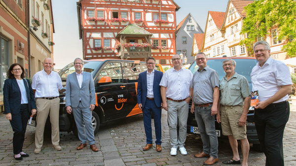 Eine Gruppe von acht Personen steht auf einer Pflasterstraße vor einem Fahrzeug mit dem Logo "vvs Rider". Im Hintergrund ist ein historisches Gebäude zu sehen. Die Personen lächeln und scheinen an einem Gemeinschaftsprojekt oder einer Veranstaltung teilzunehmen.