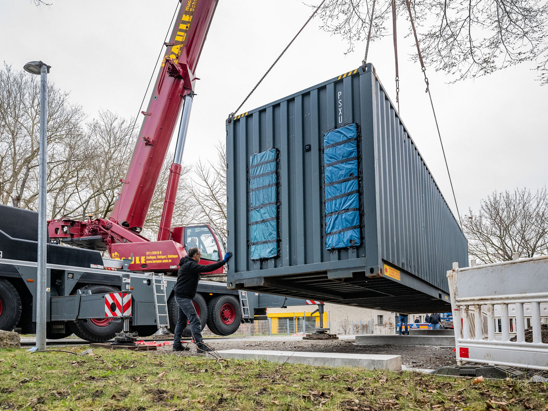 Un camion-grue soulève un conteneur métallique sur un chantier. Un travailleur supervise l'opération, tandis que le conteneur est en cours de levage. Des arbres dépouillés et des barrières de sécurité sont visibles en arrière-plan.