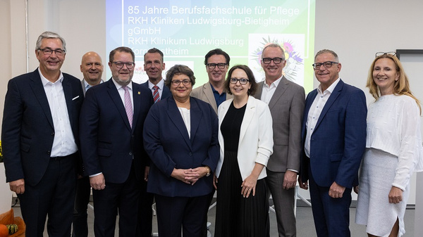 A diverse group of twelve individuals poses together for a photo, celebrating the 85th anniversary of a nursing vocational school. They stand in a well-lit room, dressed in formal attire, with a presentation screen displaying the anniversary details in the background.