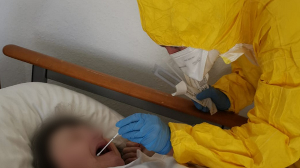 A healthcare worker in a yellow protective suit and gloves is administering a test to a patient lying in bed, covered with a white blanket. The patient has their mouth open, while the healthcare worker uses a swab for the procedure.