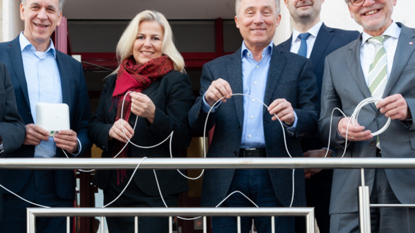 Gruppo di sei persone in piedi su un balcone, sorridenti mentre tengono in mano dei cavi. Indossano abiti formali e si trovano davanti a una porta. L'atmosfera è di festeggiamento, suggerendo una cerimonia o un evento speciale.