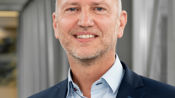 A middle-aged man with a bald head smiles while wearing a dark suit over a light blue shirt. He stands in an indoor setting with modern architecture in the background. The photo conveys a professional and confident demeanor.