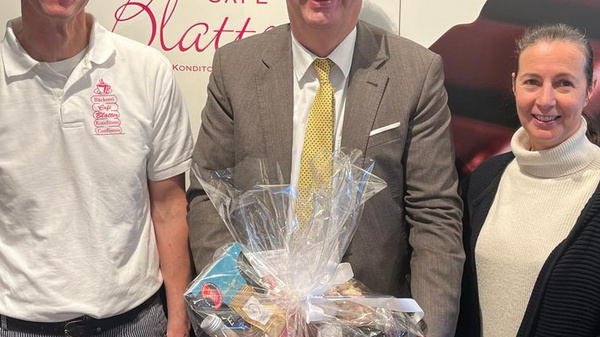 Drei Personen stehen vor einer Wand mit Schokoladendesign. In der Mitte hält ein Mann im Anzug einen Geschenkkorb mit verschiedenen Leckereien. Links steht ein Mann in einem weißen T-Shirt, rechts eine Frau in einem schwarzen Pullover. Alle lächeln in die Kamera.