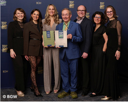 Eine Gruppe von sieben Menschen steht vor einem blauen Hintergrund mit Schriftzug "TOP 100". Sie halten Auszeichnungen in den Händen. Die Personen sind verschieden gekleidet, einige tragen formelle Kleidung. Die Atmosphäre wirkt feierlich. (Text KI generiert)