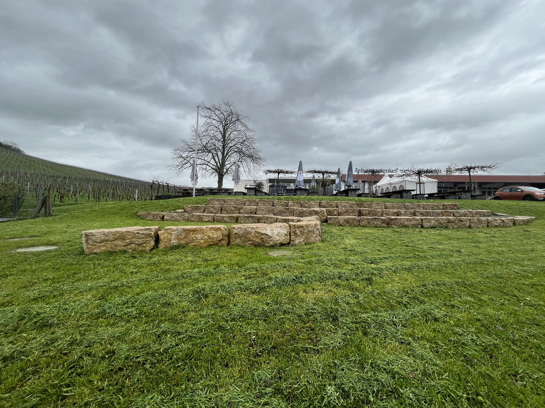 Eine kreisförmige Steinformation liegt auf einer grünen Wiese, umgeben von einem bewölkten Himmel. Im Hintergrund sind Weinreben und einige Gebäude zu sehen. Ein einzelner Baum steht in der Nähe der Steine.