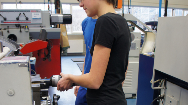Eine junge Person in schwarzem T-Shirt und Jeans steht an einer Maschinenbedieneinheit in einer Werkstatt. Sie konzentriert sich auf die Bedienung der Maschine, während eine andere Person im Hintergrund sichtbar ist. Die Umgebung deutet auf einen Ausbildungs- oder Fertigungsbereich hin.