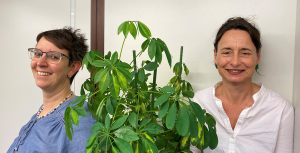 Zwei Frauen stehen nebeneinander, eine mit Brille und kurz geschnittenen Haaren, die andere mit langen Haaren. Zwischen ihnen befindet sich eine grüne Pflanze. Beide lächeln freundlich und tragen legere Kleidung. Der Hintergrund ist hell und neutral gehalten.
