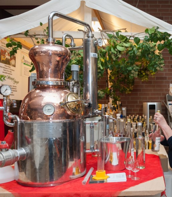 Eine große Destillationsanlage aus Kupfer und Edelstahl steht in einem festlich dekorierten Raum. Umgeben von Flaschen und Gläsern zeigt sie den Prozess der Spirituosenherstellung. Im Hintergrund sind grüne Pflanzen sichtbar, die zur Atmosphäre beitragen.