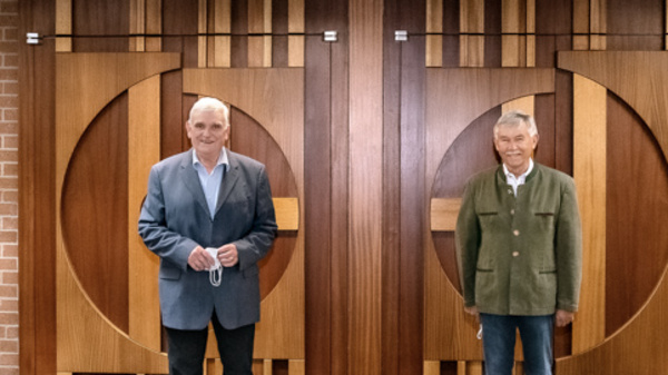 Four men stand in a conference room, each positioned in front of large wooden doors. They are dressed in formal and semi-formal attire, and they are looking towards the camera, with neutral expressions. The setting features warm wood tones and brick walls.