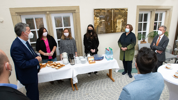Un groupe de personnes se tient debout autour d'une table avec des boissons et des collations. Elles portent toutes des masques faciaux. Un homme, probablement en train de parler, est en face des autres participants dans un cadre chaleureux et accueillant.