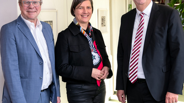 Drei Menschen stehen in einem hellen Raum, alle lächeln und blicken zur Kamera. Sie tragen formelle Kleidung. Im Hintergrund sind einige Bilder an der Wand und eine Pflanze sichtbar.