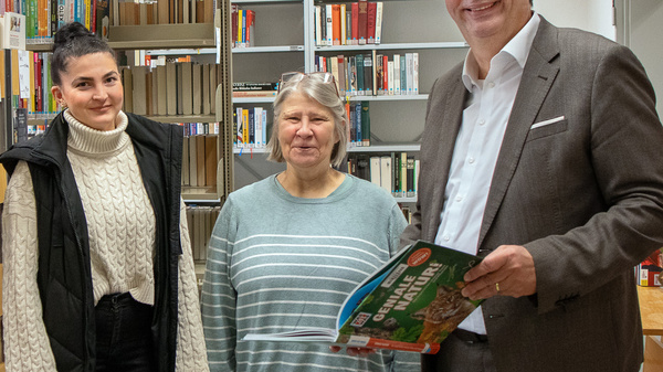 Vier Personen stehen in einer Bibliothek zwischen Bücherregalen. Der Mann rechts hält ein Buch in der Hand. Die anderen Personen lächeln und stehen nebeneinander, während sie freundlich in die Kamera blicken. Die Atmosphäre wirkt einladend und gemeinschaftlich.