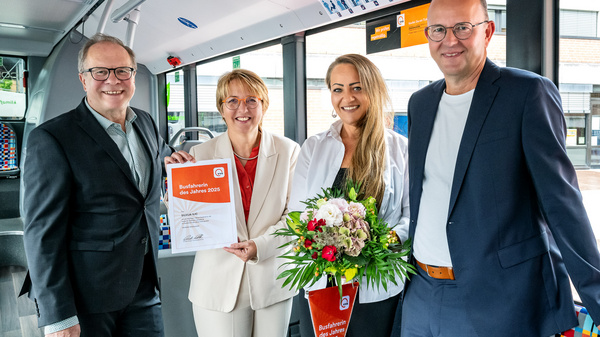 Vier Personen stehen in einem Bus und lächeln. Eine Frau hält ein Zertifikat in der Hand, während eine andere einen Blumenstrauß präsentiert. Alle drei sind elegant gekleidet. Im Hintergrund sind Busfenster und Sitzpolster sichtbar.