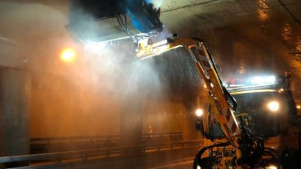 Ein Reinigungsfahrzeug reinigt unter einer Brücke. Hochdruckwasser wird eingesetzt, um den Beton von Schmutz zu befreien. Die Umgebung ist nass, und die Straßenbeleuchtung strahlt in die dunkle Szenerie.