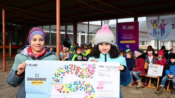 Zwei Kinder stehen vor einer Gruppe von Schülern und halten ein buntes Plakat mit einem Wegdiagramm in den Händen. Im Hintergrund sind einige Stühle und Banner mit dem Text „Schulwegprofis“ zu sehen. Der Ort wirkt festlich und lehrreich.