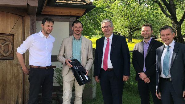 Fünf Männer stehen in einer grünen Landschaft vor einem Holzhaus. Einer von ihnen hält ein schwarzes Objekt in der Hand. Alle tragen formelle Kleidung und lächeln. Die Szenerie vermittelt einen positiven, festlichen Eindruck.