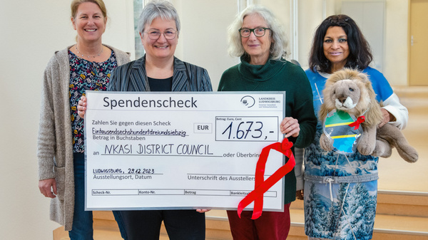 Vier Frauen stehen zusammen und halten einen großen Scheck mit dem Betrag von 1.673 Euro, der an den Nkasi District Council übergeben wird. Die Frauen lächeln und eine von ihnen hält ein Stofftier. Der Hintergrund ist hell und freundlich.