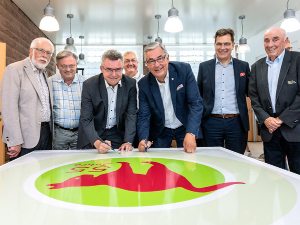 20.07.2025 - 55 Jahre Schullandheim Stümpfelbrunn Eine Gruppe von acht Männern steht hinter einem großen Tisch, auf dem ein Logo mit einem roten Tier und einem grünen Hintergrund zu sehen ist. Sie lächeln und einige von ihnen halten Stifte, während sie gemeinsam an dem Tisch stehen.
