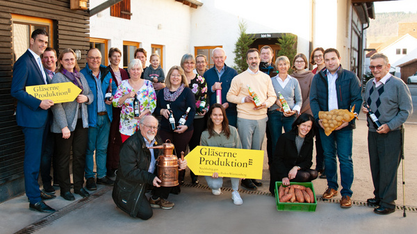 A diverse group of people stands together outside a building, holding various food items and signs. One sign reads "Gläserne Produktion," indicating a connection to transparent production or local food. The gathering suggests a celebration or community event focused on local produce.