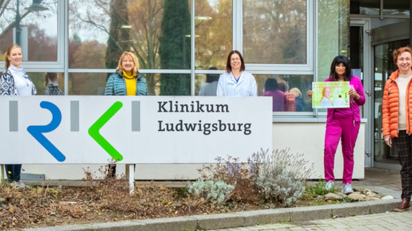 Des femmes de différents âges et styles se tiennent devant le Klinikum Ludwigsburg, tenant des affiches. Elles portent des vêtements colorés et affichent des sourires, symbolisant la solidarité et le soutien. Un panneau du Klinikum est clairement visible en arrière-plan.