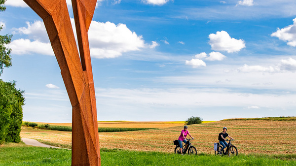 Die Aufnahme zeigt eine große, abstrakte Skulptur aus Metall, die auf einer Wiese steht. Im Hintergrund verlaufen ein Weg und Felder. Zwei Radfahrer fahren in der Nähe der Skulptur, umgeben von einer üppigen, grünen Landschaft und einem strahlend blauen Himmel mit Wolken.