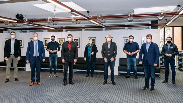 Eine Gruppe von neun Personen steht in einem modernen, hellen Raum. Alle tragen Mund-Nasen-Schutz. Die Personen sind formell gekleidet und stehen in zwei Reihen hintereinander. Im Hintergrund sind einige Gemälde an den Wänden sichtbar. (Text KI generiert)