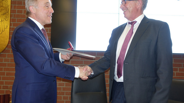 Two men are engaged in a handshake, with one presenting a document to the other. They appear to be in an official setting with a screen in the background displaying the name "Landkreis Ludwigslust." The atmosphere suggests a formal meeting or signing ceremony.