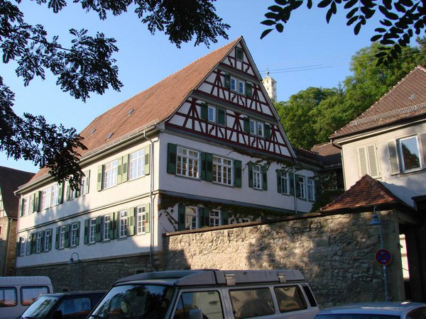 Ein historisches Haus mit Fachwerkarchitektur, das mehrere Fenster mit grünen shutters hat. Das Dach ist rot gedeckt und es gibt einige Bäume im Vordergrund. Im Hintergrund sind weitere Gebäude sichtbar.