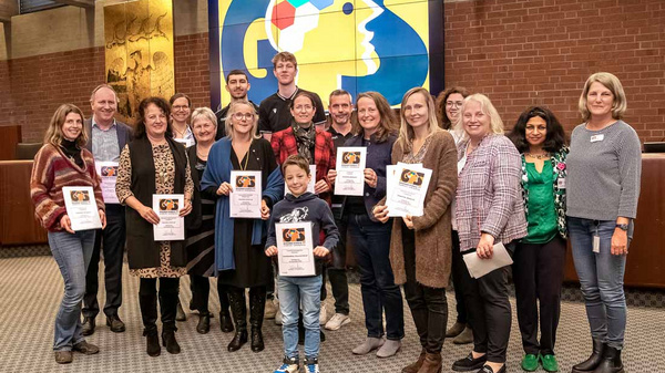 A diverse group of 16 people stands together, holding certificates in a spacious room. There are men, women, and a child, all smiling. In the background, a colorful logo is displayed on a screen, suggesting a celebratory event or awards ceremony.