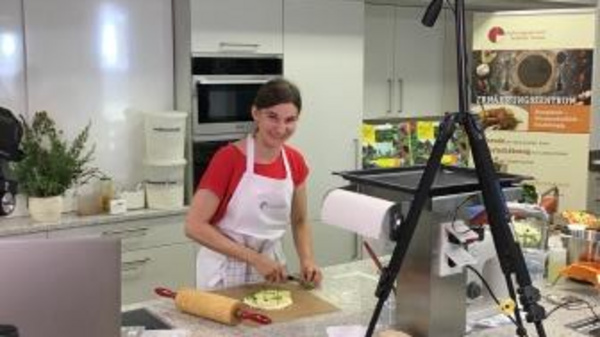A person wearing an apron smiles while preparing food in a modern kitchen. Ingredients and cooking tools are arranged on a countertop, with a camera set up above to capture the cooking process. The kitchen has a clean, organized appearance.