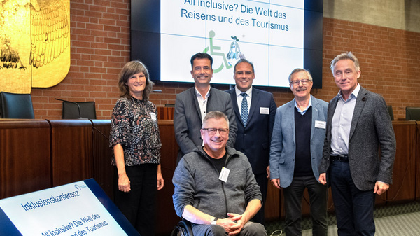 A group of six individuals, including one person in a wheelchair, pose for a photo at an inclusion conference focused on accessible travel and tourism. A presentation screen in the background displays the conference title in German.