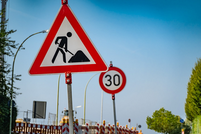 Das Bild zeigt ein Verkehrsschild mit der Warnung vor Baustellen, dargestellt durch eine Person, die mit einer Schaufel arbeitet. Neben dem Baustellenschild ist ein weiteres Schild mit einer Geschwindigkeitsbegrenzung von 30 km/h zu sehen.